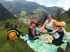 Mehrere Kinder und eine erwachsene Frau sitzen auf einer grünen Picknickdecke auf einem Berg, im Hintergrund sieht man mehrere Berge