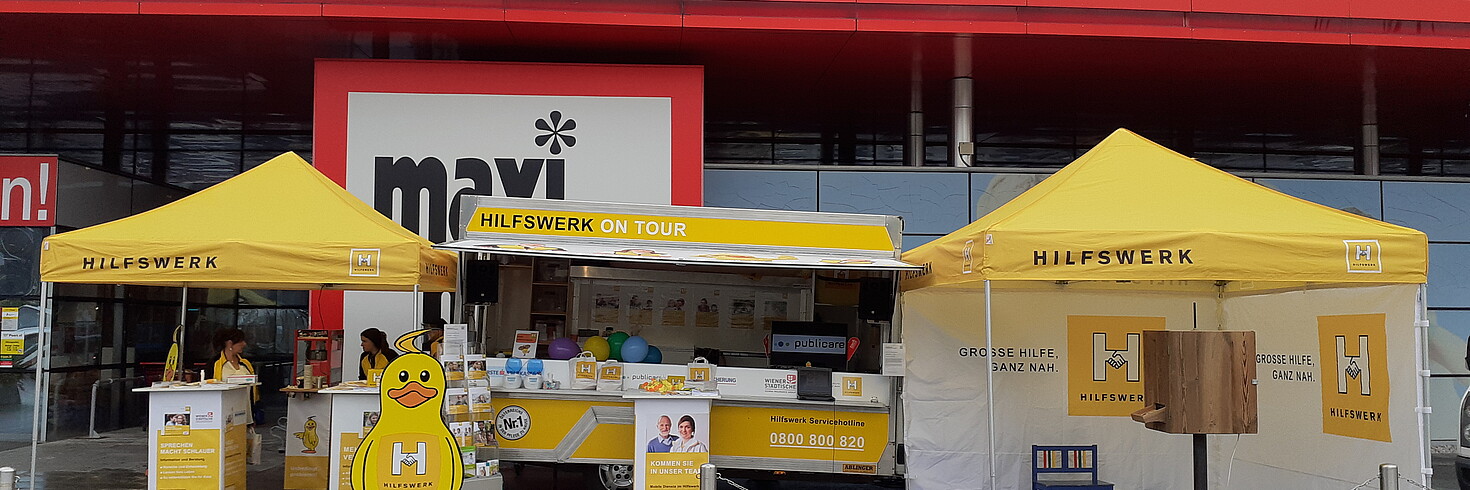 Hilfswerk Salzburg Hilfswerk on Tour steht vor Maximarkt, Stände und Schirme sind zu sehen, Infoveranstaltung und Beratung 