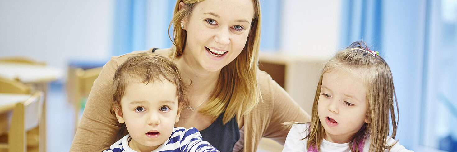 Eine Pädagogin sitzt zwischen zwei Kindern und schaut in die Kamera