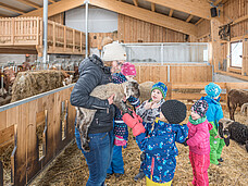 Mehrere Kinder stehen im Stall, eine erwachsene Frau hält ein Schaf