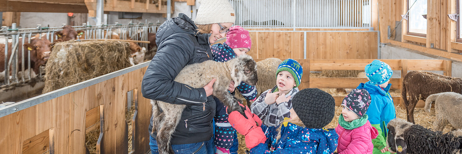 Mehrere Kinder stehen im Stall, eine erwachsene Frau hält ein Schaf
