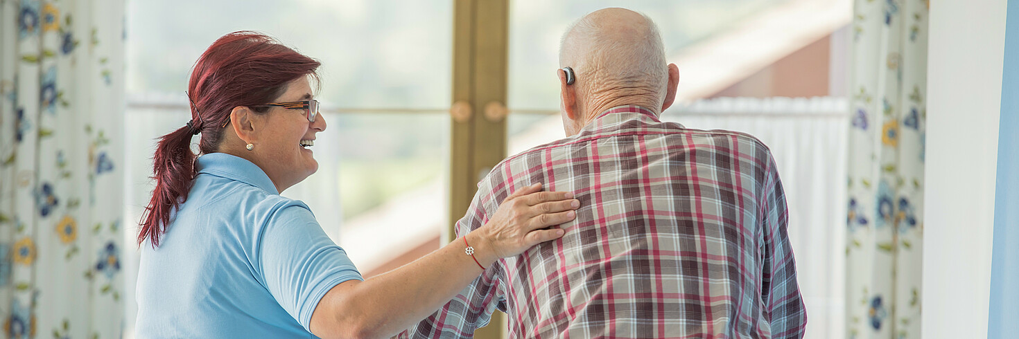 Eine Dame und ein älterer Herr spazieren gemeinsam, die Frau berührt den Mann an der Schulter, der Mann hat einen Rollator