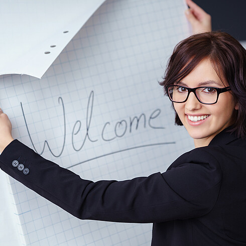 Frau steht vor Flipchart mit Text "Welcome" und lächelt in die Kamera.