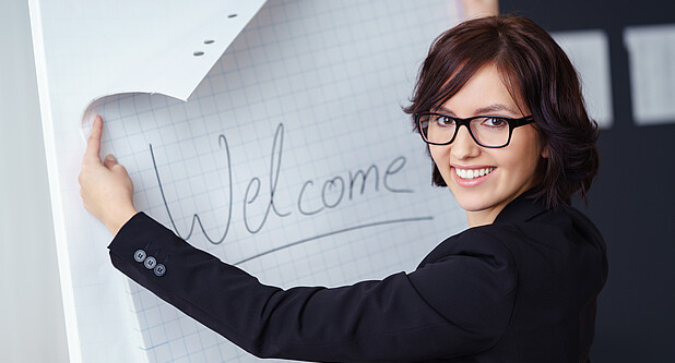Frau steht vor Flipchart mit Text "Welcome" und lächelt in die Kamera.