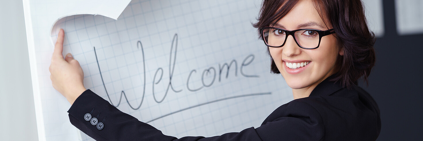 Frau steht vor Flipchart mit Text "Welcome" und lächelt in die Kamera.