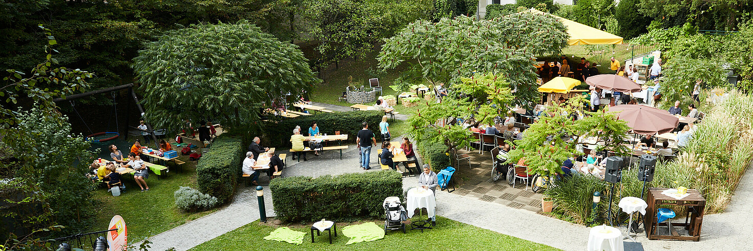 Mehrere Menschen feiern in einem großen Garten ein sommerliches Fest. Es gibt eine kleine Bühne und die Leute sitzen gemeinsam an Tischen.