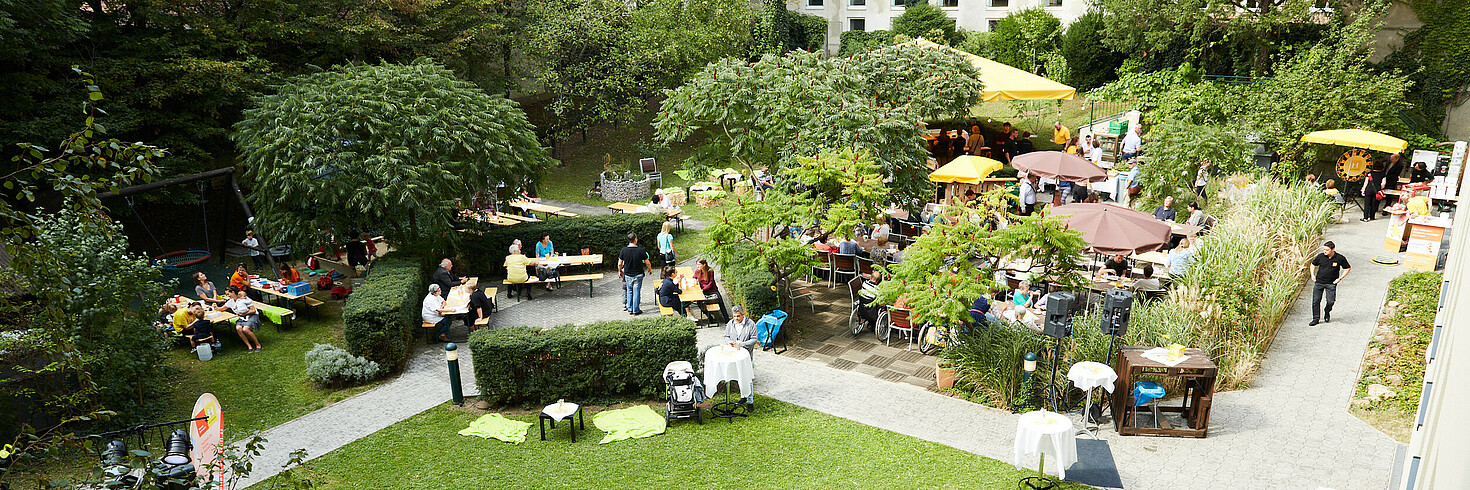 Mehrere Menschen feiern in einem großen Garten ein sommerliches Fest. Es gibt eine kleine Bühne und die Leute sitzen gemeinsam an Tischen.