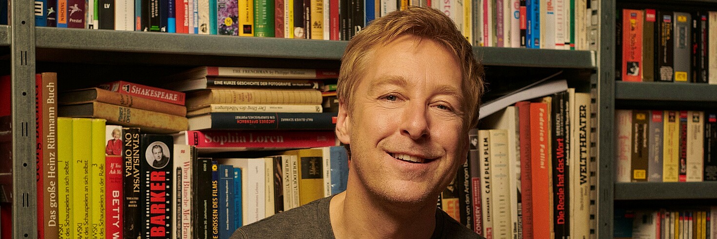 Bild von einem Mann mit blonden kurzen Haaren vor einem Bücherregal