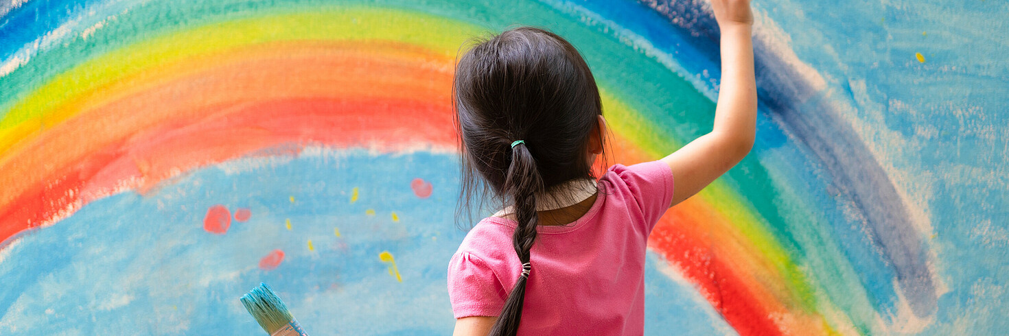Mädchen von hinten mit dunklen Haaren und Zopf. Zeichnet einen Regenbogen.
