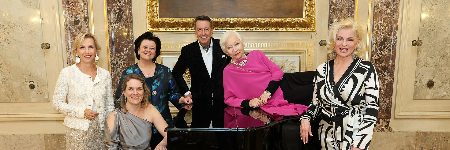 Fünf Frauen und ein Mann festlich gekleidet bei einem Klavier
