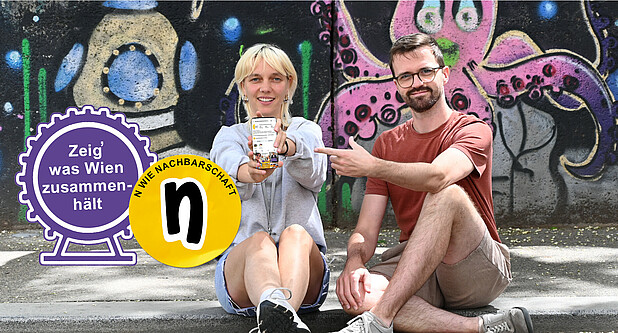 Eine Frau und ein Mann sitzen vor einem Graffiti und zeigen auf ein Handy
