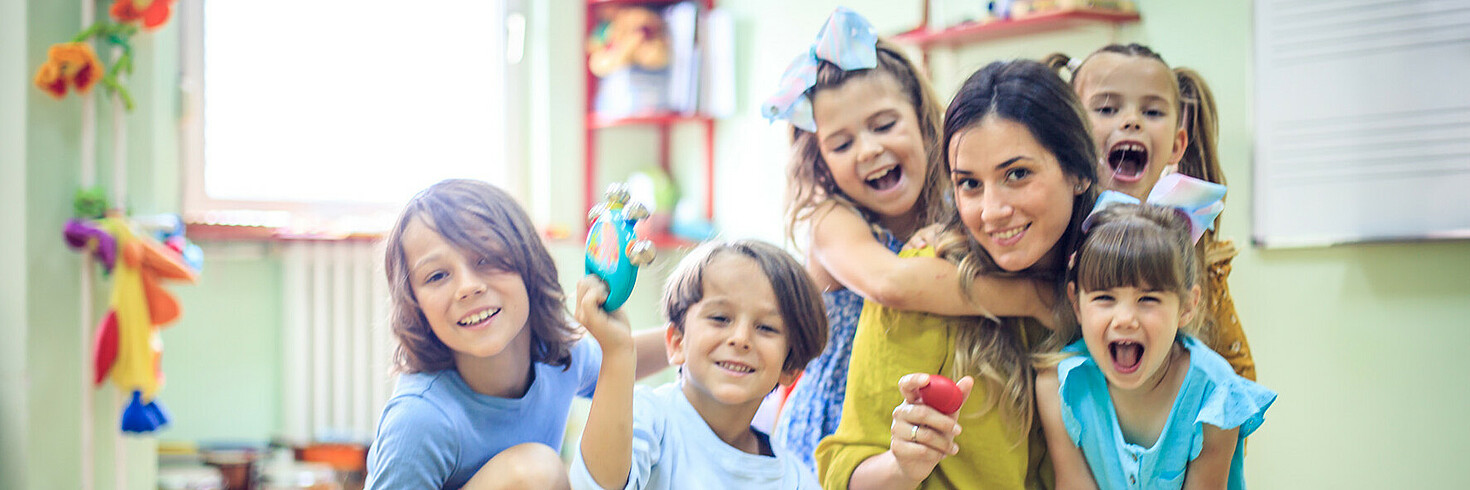 Frau sitzt mit fünf Kindern am Boden in einer Kindergartengruppe