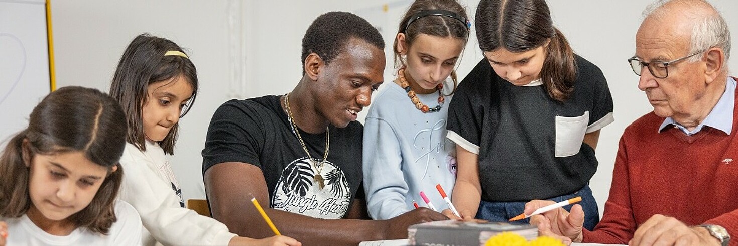 Zwei Erwachsene Männer sitzen mit Kindern am Tisch und helfen ihnen beim Lernen.